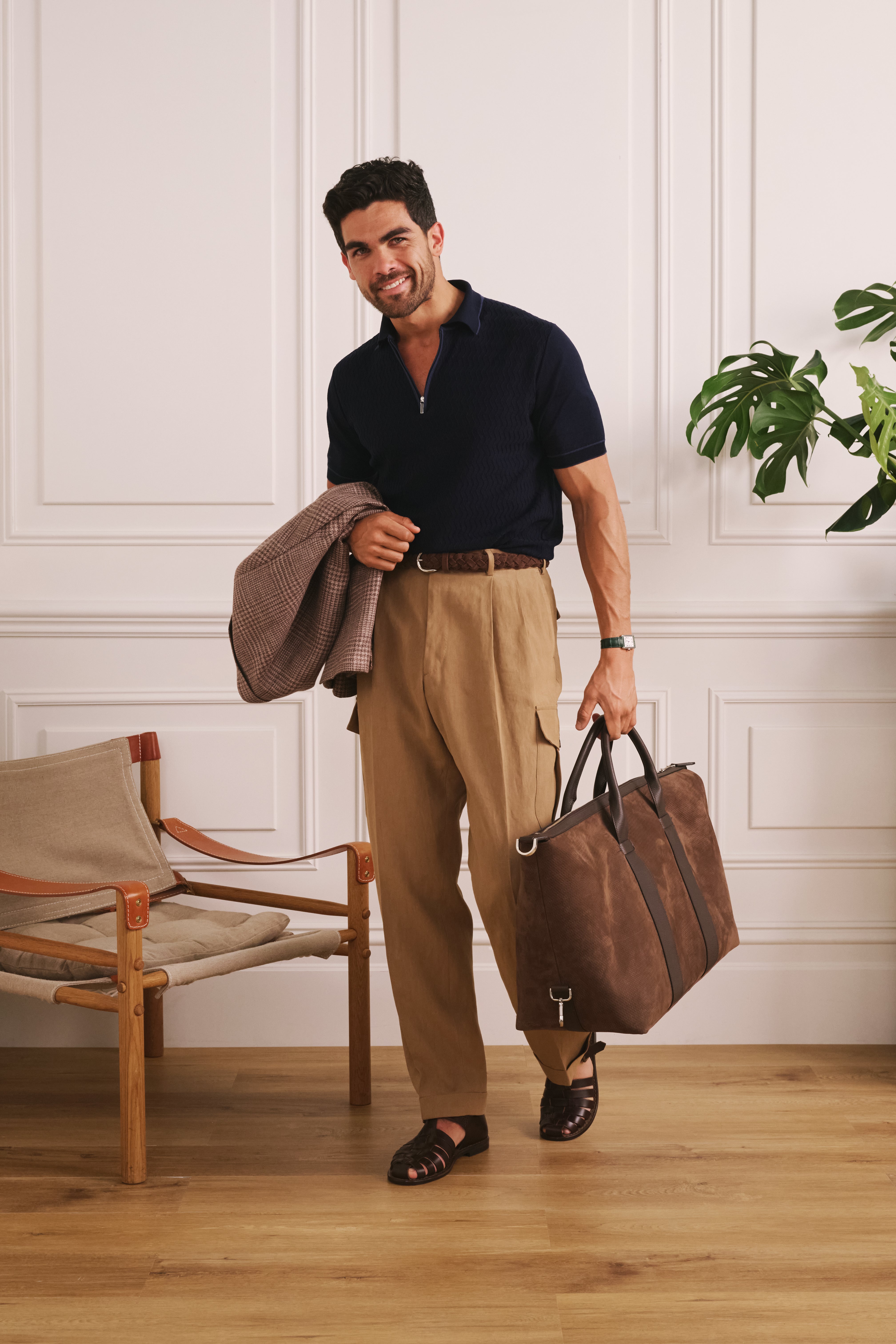 Man holding a brown leather bag in a room with wooden floor and white paneled walls.