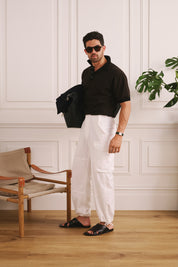 Man in black polo shirt and white pants standing in a room with wooden floor and white walls.