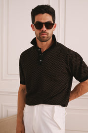Man wearing a black patterned polo shirt and white pants against a white wall.