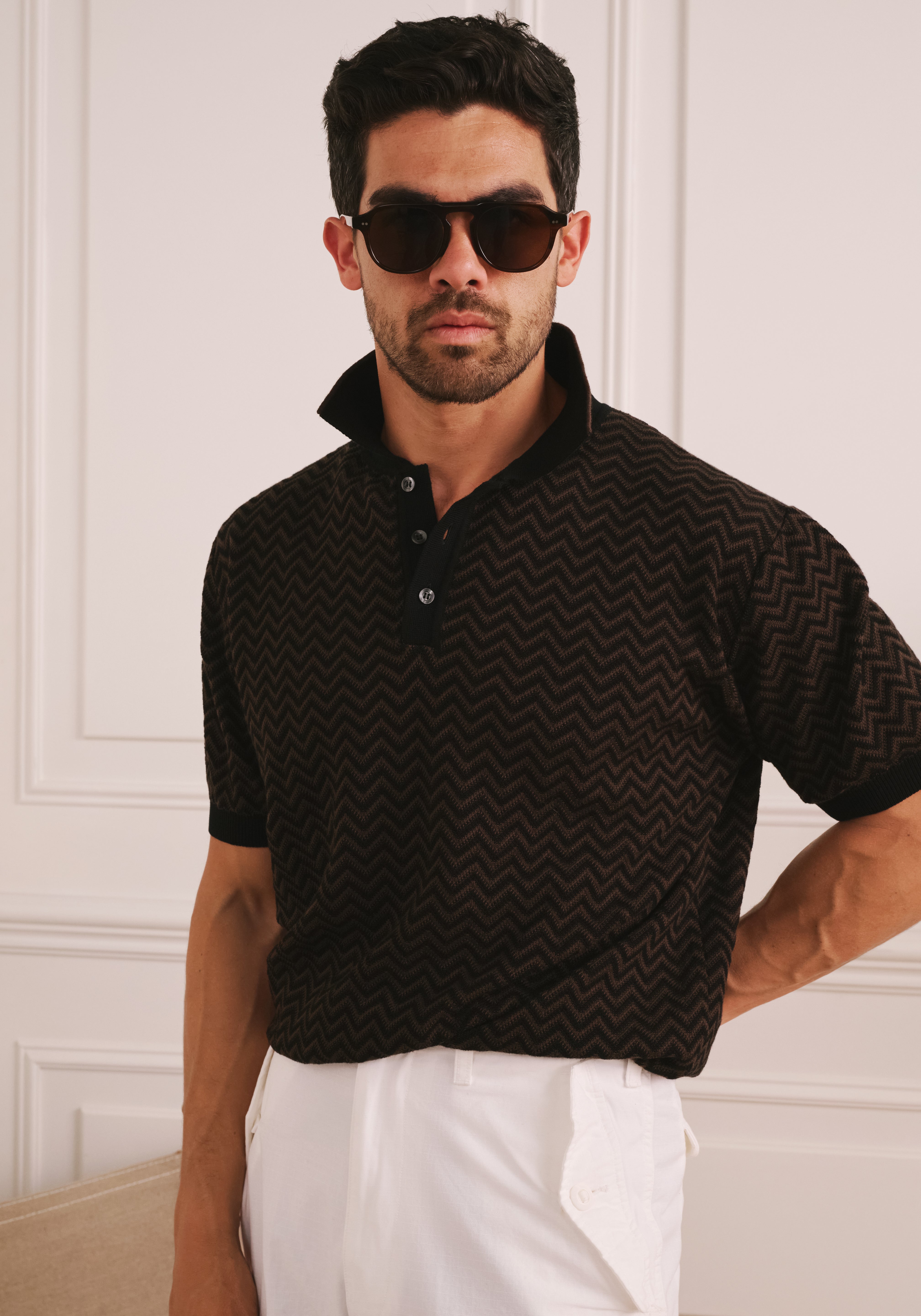 Man wearing a black patterned polo shirt and white pants against a white wall.