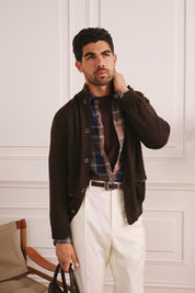 Man wearing a brown jacket, plaid shirt, and white pants against a white paneled wall.