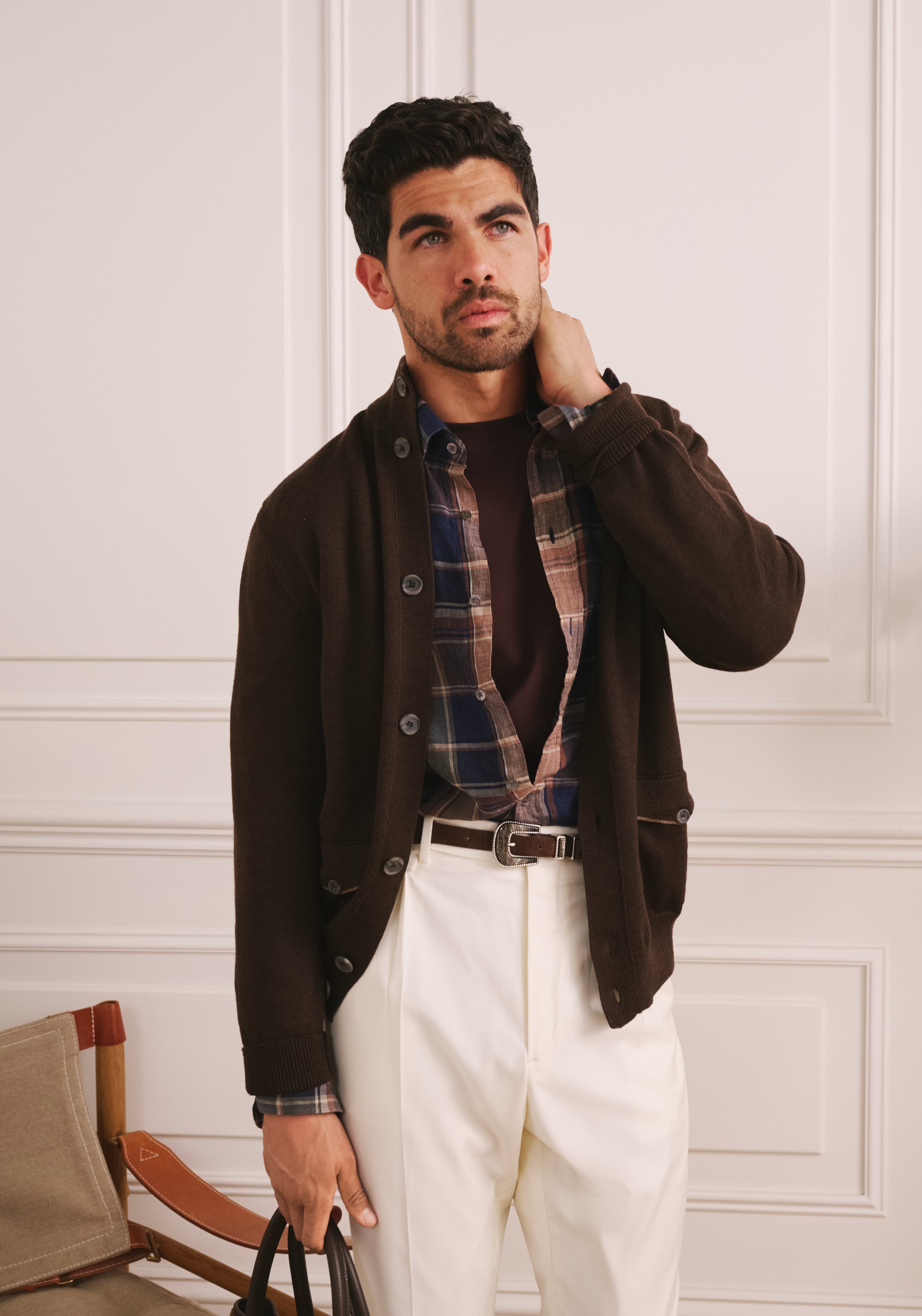 Man wearing a brown jacket, plaid shirt, and white pants against a white paneled wall.