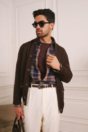 Man wearing sunglasses, a brown jacket, plaid shirt, and white pants against a white paneled wall.