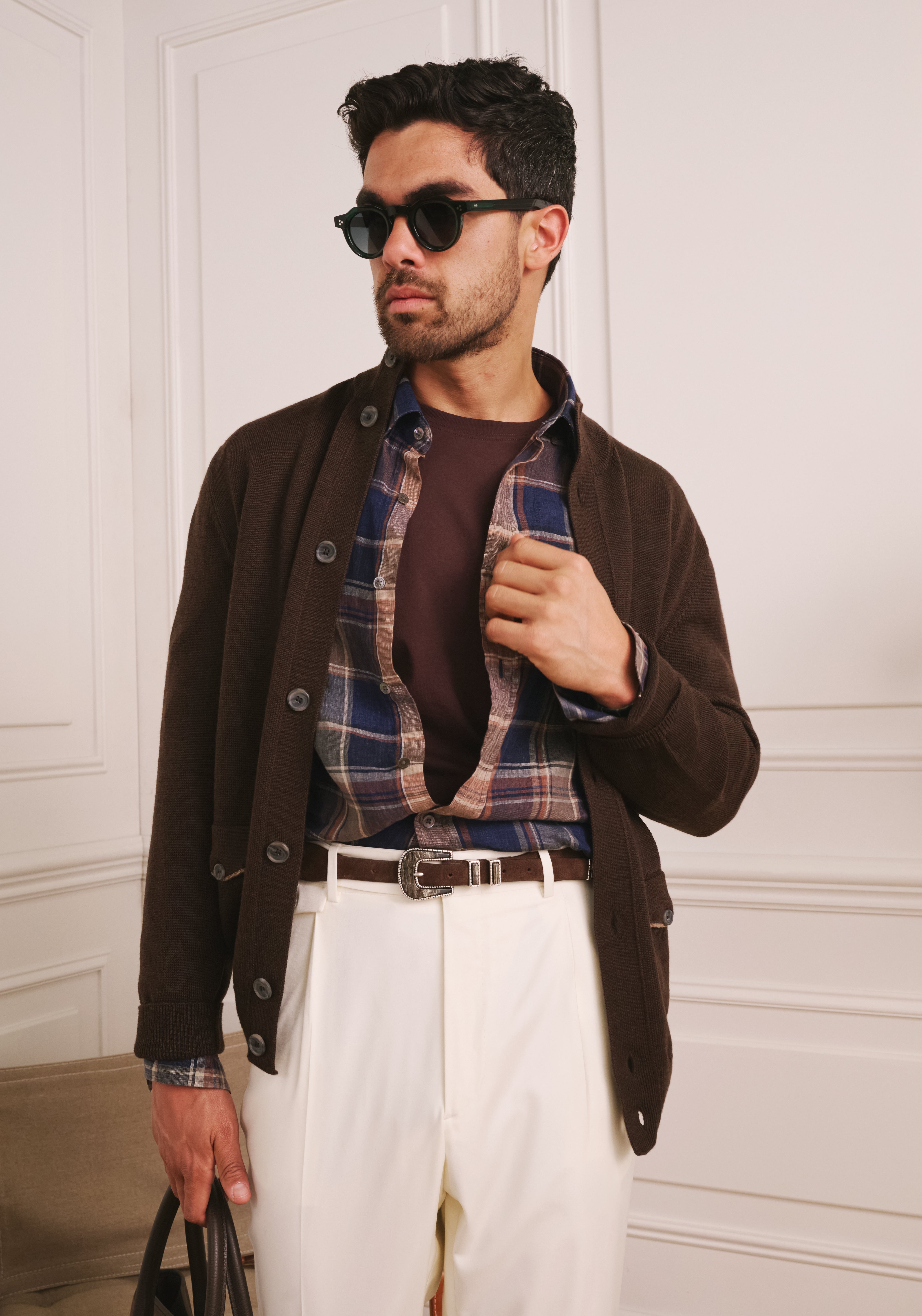 Man wearing sunglasses, a brown jacket, plaid shirt, and white pants against a white paneled wall.