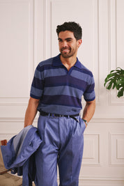 Man wearing a blue striped polo shirt and pants, holding a jacket indoors.