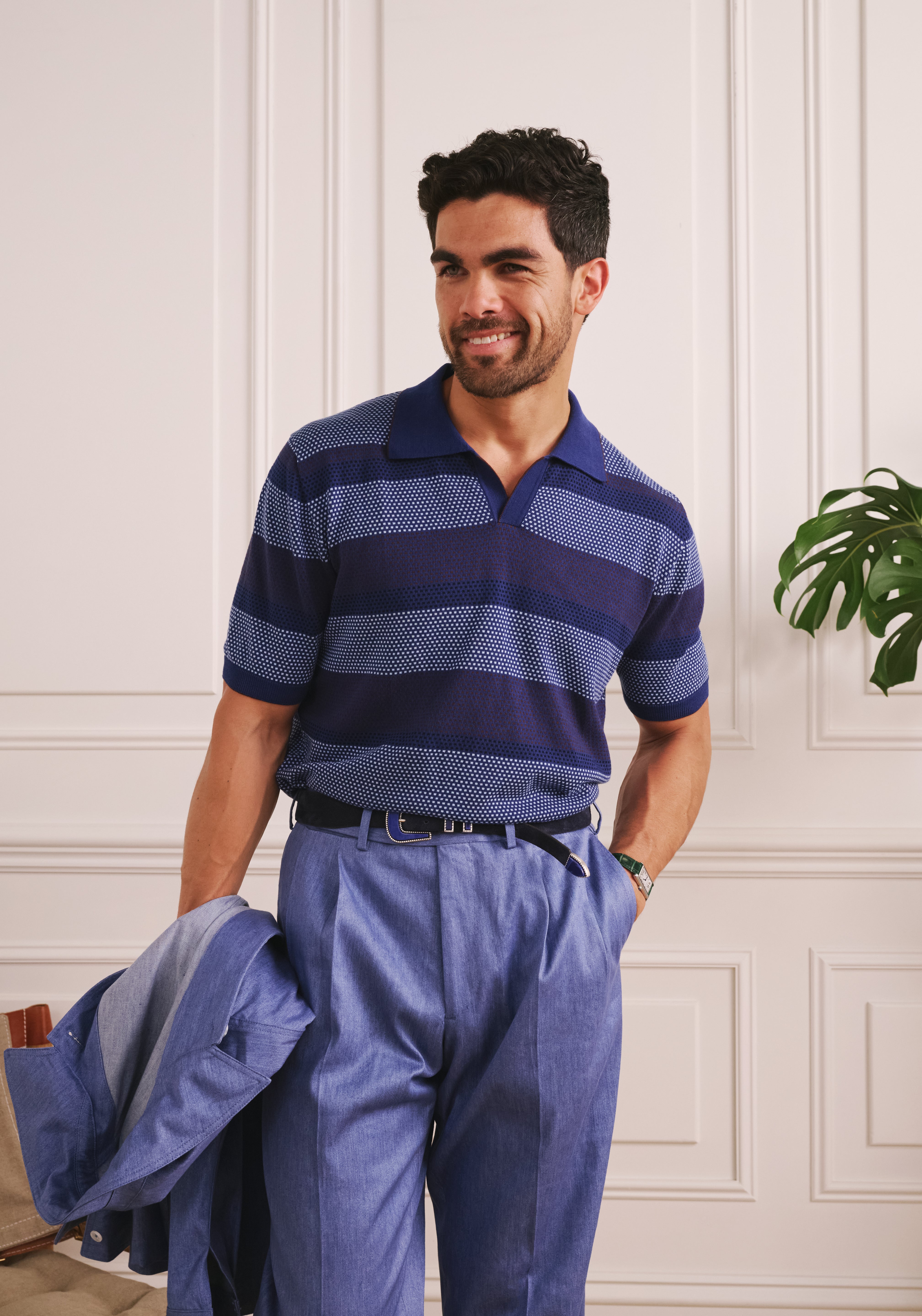 Man wearing a blue striped polo shirt and pants, holding a jacket indoors.