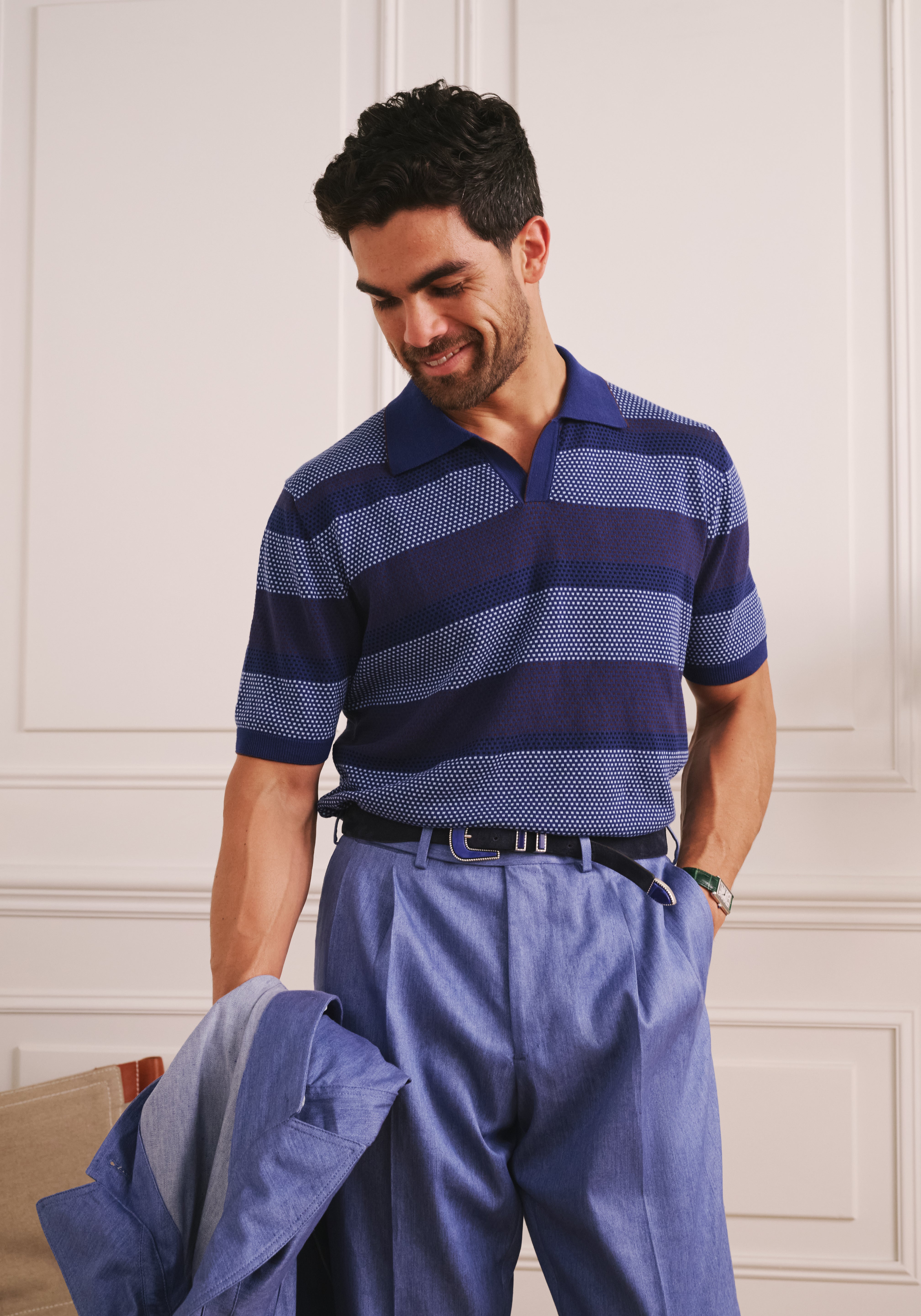 Man wearing a blue and navy striped polo shirt and pants, standing against a white paneled wall.