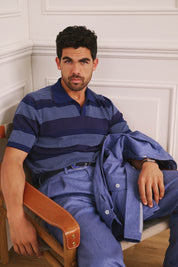 Man sitting on a chair wearing a blue striped shirt and blue pants with a white wall background