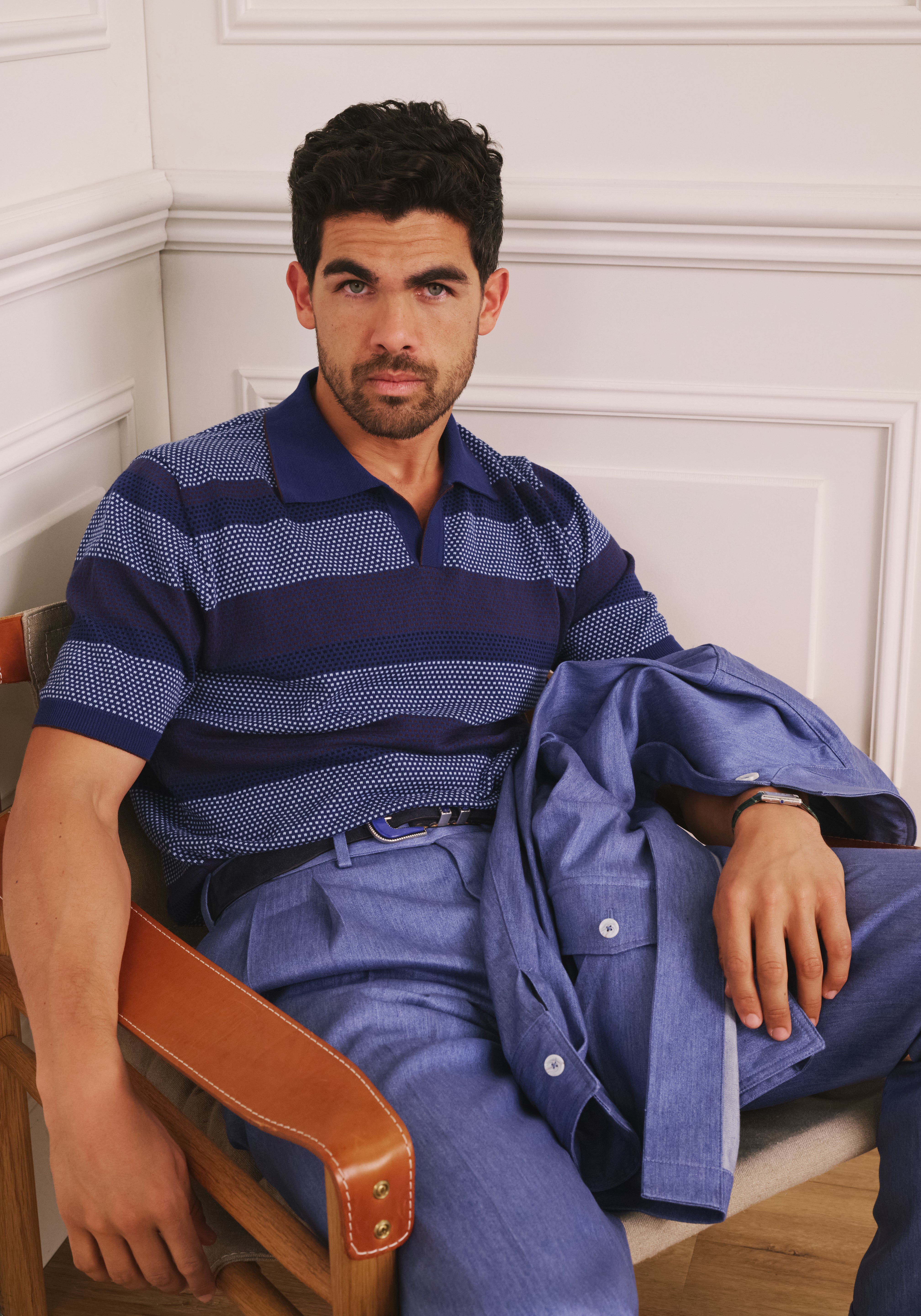 Man sitting on a chair wearing a blue striped shirt and blue pants with a white wall background