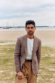 Man standing on a beach wearing a brown blazer and beige pants.