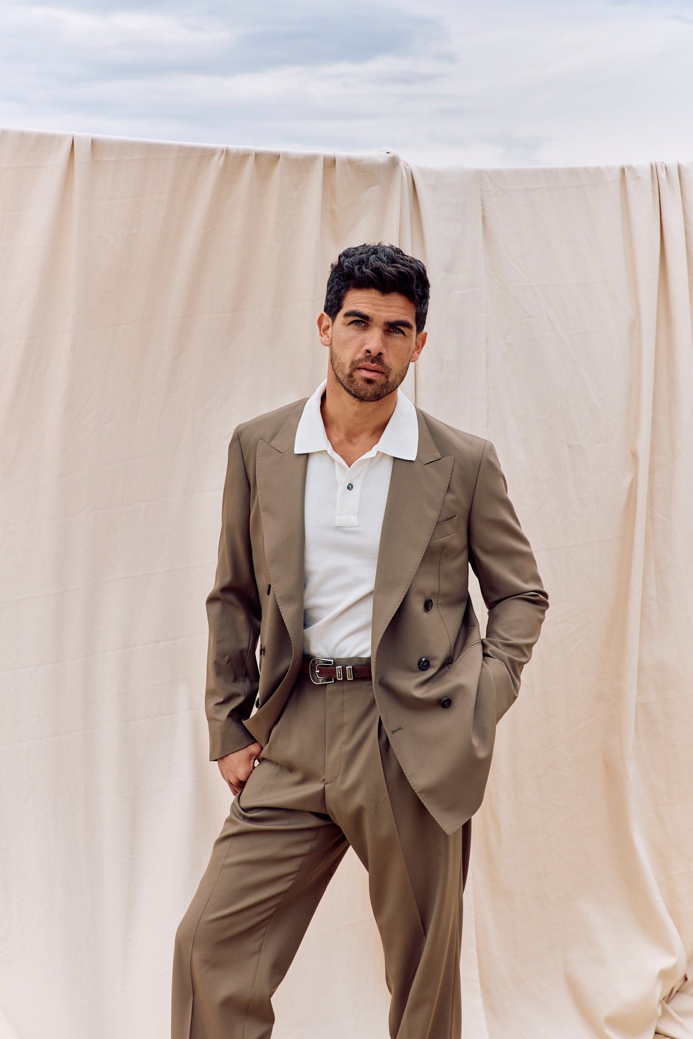 Man wearing a beige suit standing against a beige curtain with a blue sky background