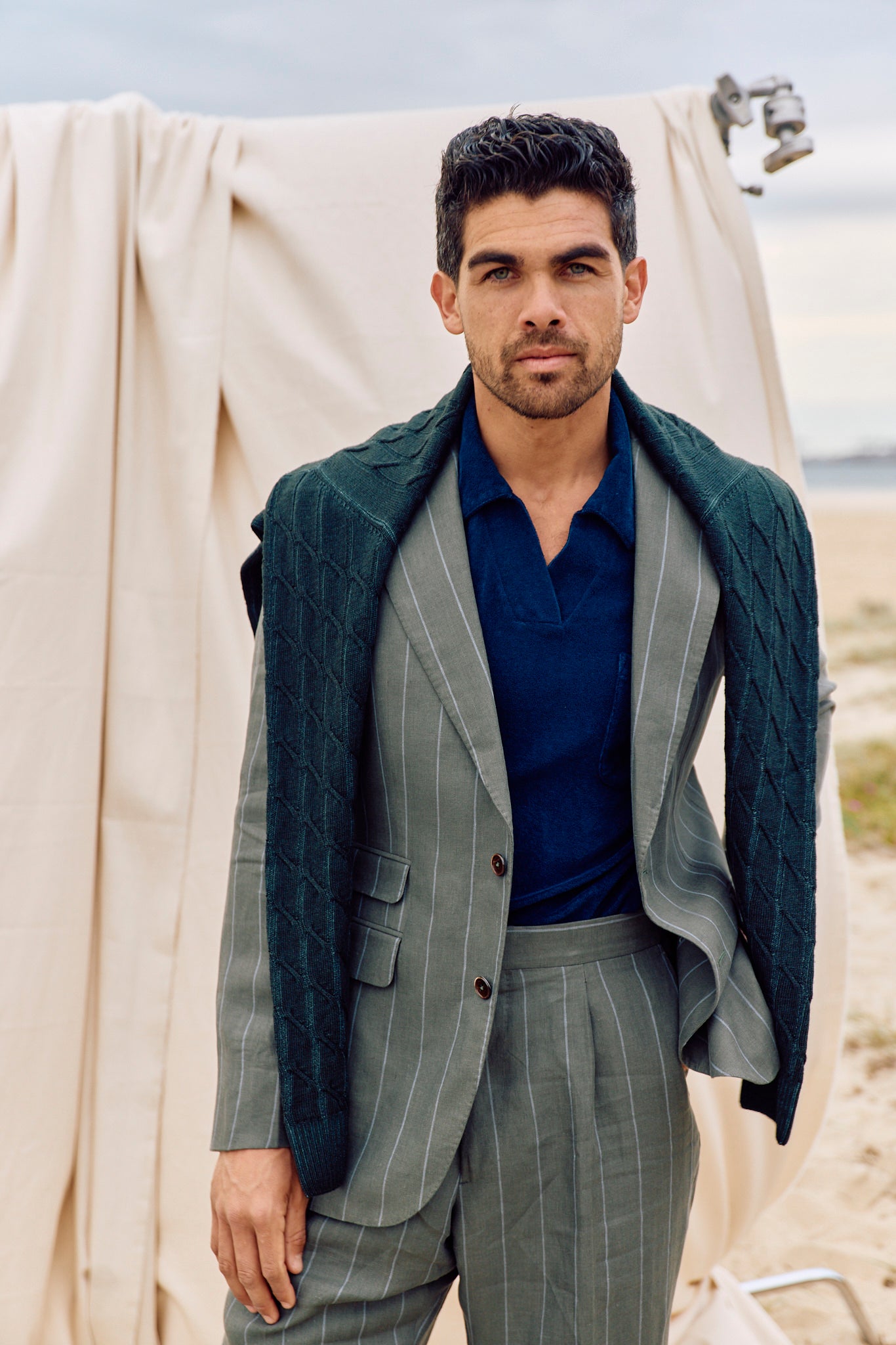 Man wearing a gray suit with a blue shirt and cable knit cardigan against a neutral background